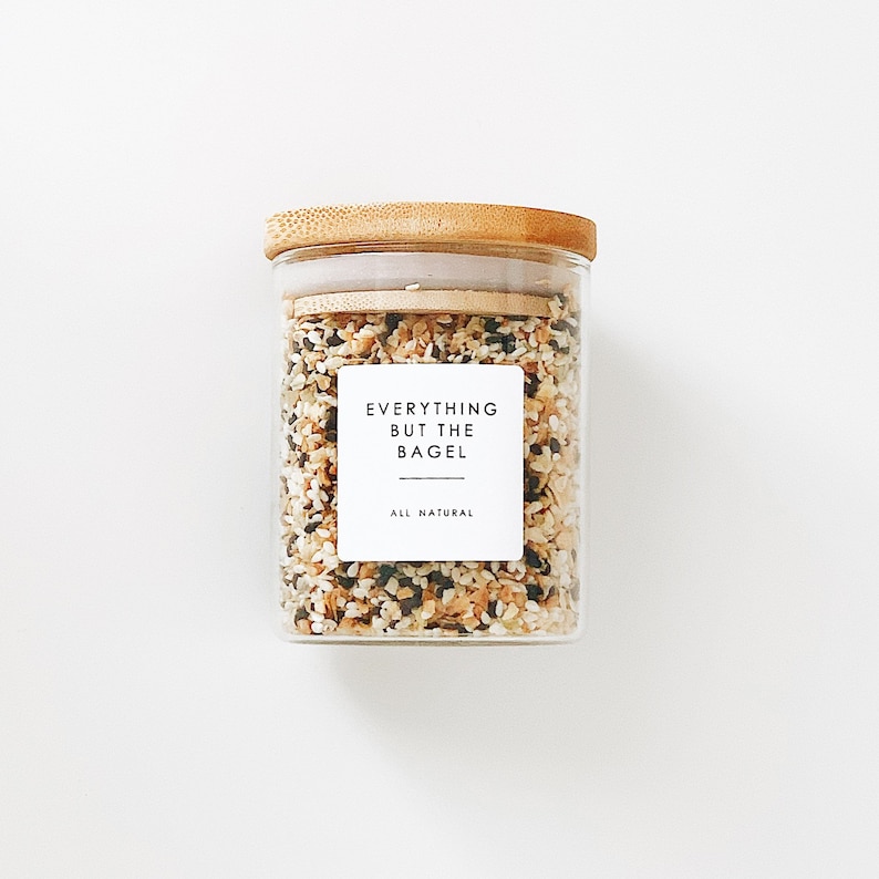 May include: A glass jar with a bamboo lid filled with a blend of sesame seeds, poppy seeds, dried onion, and garlic. The jar has a white label that reads "Everything But The Bagel" and "All Natural".
