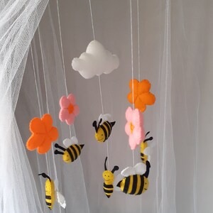 May include: A handmade baby mobile featuring felt bees, flowers, and a cloud. The bees are yellow and black, the flowers are pink and orange, and the cloud is white. The mobile is suspended from a wooden frame.