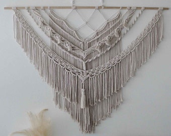 Pode incluir: Um grande pendurado de parede de macramé de cor branco-queimado com um design geométrico e franjas. O pendurado é feito de fibras naturais e tem um estilo boémio.
