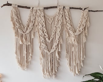 Pode incluir: Uma tapeçaria de macramé de parede com três seções, cada uma com um padrão diferente. A tapeçaria é feita de cordão de cor natural e está presa a um galho. A tapeçaria é exibida em uma parede branca com uma planta em vaso no canto.