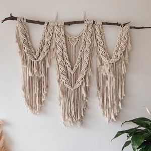 May include: A macrame wall hanging with three sections, each featuring a different pattern. The hanging is made of natural-coloured cord and is attached to a branch. The hanging is displayed on a white wall with a potted plant in the corner.