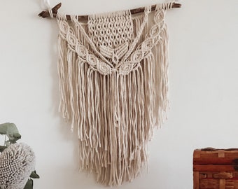 Puede incluir: Un tapiz de macramé beige con una rama de madera en la parte superior y flecos en la parte inferior. El tapiz está frente a una pared blanca y hay cajas de madera apiladas sobre una mesa de madera en primer plano.