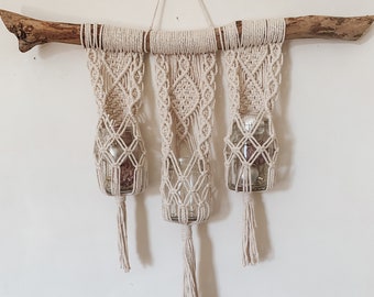 May include: A macrame wall hanging with three glass jars, each with a tassel. The wall hanging is made of natural beige cotton cord and is hung from a branch.