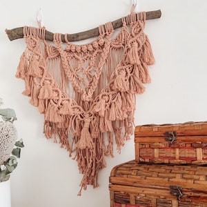 May include: A macrame wall hanging with a natural wood branch and tassels in a light peach color. The hanging is displayed on a white wall with three wooden boxes stacked on a table below.