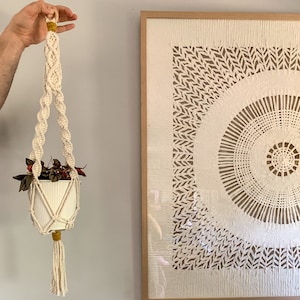 May include: A white macrame plant hanger with a yellow tassel hangs from a wooden dowel. A small white ceramic pot with a plant is inside the hanger.