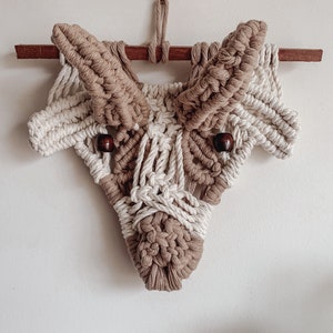 May include: A macrame wall hanging in the shape of a stylized animal head, with brown and white yarn and wooden beads for eyes.