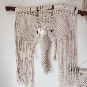 Puede incluir: Un tapiz de macramé beige en forma de cabeza de elefante con una trompa larga. El tapiz está sujeto a una rama y tiene detalles de flecos.