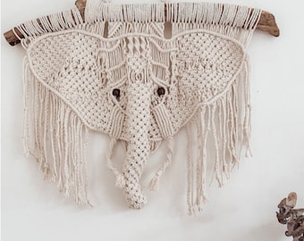 Puede incluir: Un tapiz de macramé en forma de cabeza de elefante con una rama de madera natural en la parte superior. El elefante está hecho de hilo de color crema y tiene flecos alrededor de los bordes.