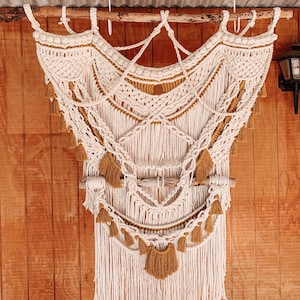May include: A white and mustard yellow macrame wall hanging with fringe. The hanging is made of knotted cords and has a wooden dowel at the top.