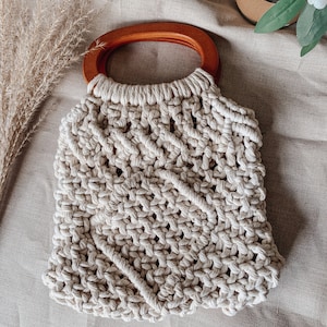 May include: A white crocheted tote bag with a brown wooden handle. The bag has a woven pattern and is made of thick rope.