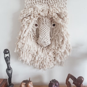 May include: A macrame wall hanging featuring a lion's face with a white background. The lion's face is made of knotted rope and has a fringe around the edges. There are three wooden animal figurines on a wooden shelf below the wall hanging.