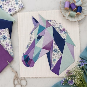 Può includere: Un pannello in tessuto trapuntato con un design geometrico a testa di cavallo composto da triangoli di tessuto in varie tonalità di viola, blu e verde acqua, intervallati da pezzi di tessuto a motivi floreali su uno sfondo color crema.