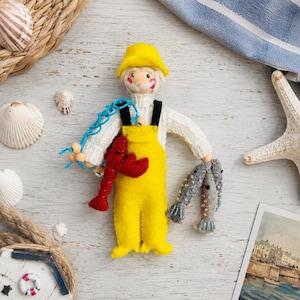May include: A felt doll of a fisherman wearing a yellow hat and yellow overalls over a white sweater. The doll holds a red lobster in one hand and two grey fish in the other, with a blue net draped over one shoulder, featuring stitched details.