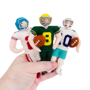 May include: Three handmade felt football player dolls. One doll wears a white jersey with red accents and the number 10, another wears a green jersey with the number 8, and the third wears a white jersey with the number 10. Each doll holds a brown football.