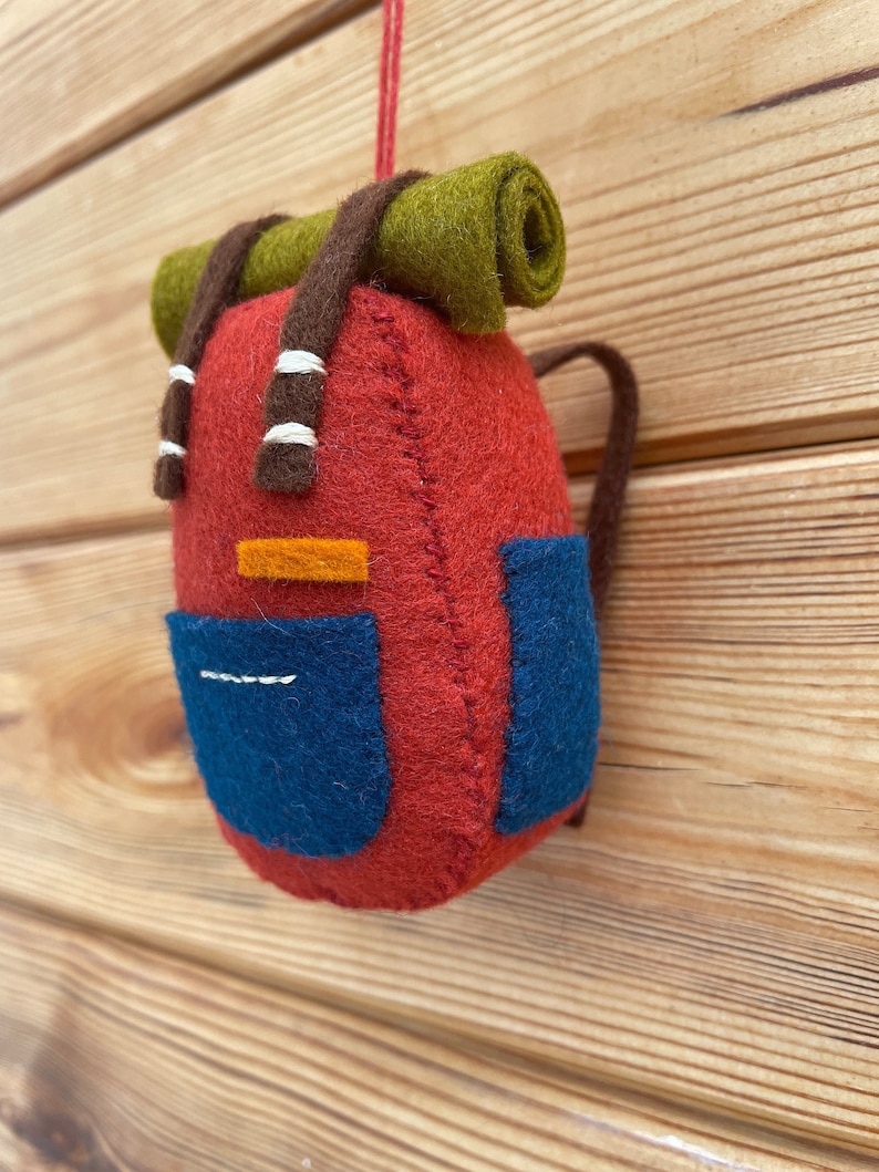 May include: A felt backpack ornament in red with blue side pockets, a green rolled sleeping bag attached to the top, and brown shoulder straps. The backpack features a small orange rectangular accent and white stitched details on the straps and pocket.