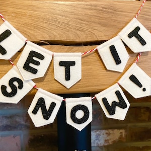 Peut inclure: Bannière en feutre blanc avec des lettres noires qui épellent "Let it snow!" accrochée à une surface en bois.