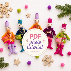 May include: Five felt nutcracker dolls in orange, blue, yellow, purple, and green uniforms with matching hats and drums. A circular white label in the center displays the text PDF photo tutorial in pink and purple script and block letters.