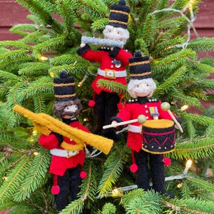 May include: Three felt Christmas ornaments depicting a marching band. The ornaments are red, black, and gold. One ornament plays a flute, one plays a drum, and one plays a trumpet.