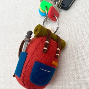 May include: A felt keyring shaped like a miniature backpack in rust red with a dark olive green bedroll on top, a blue front pocket, brown straps, and a small grey tool with three dark beads attached to the side, hanging from a metal ring with keys.