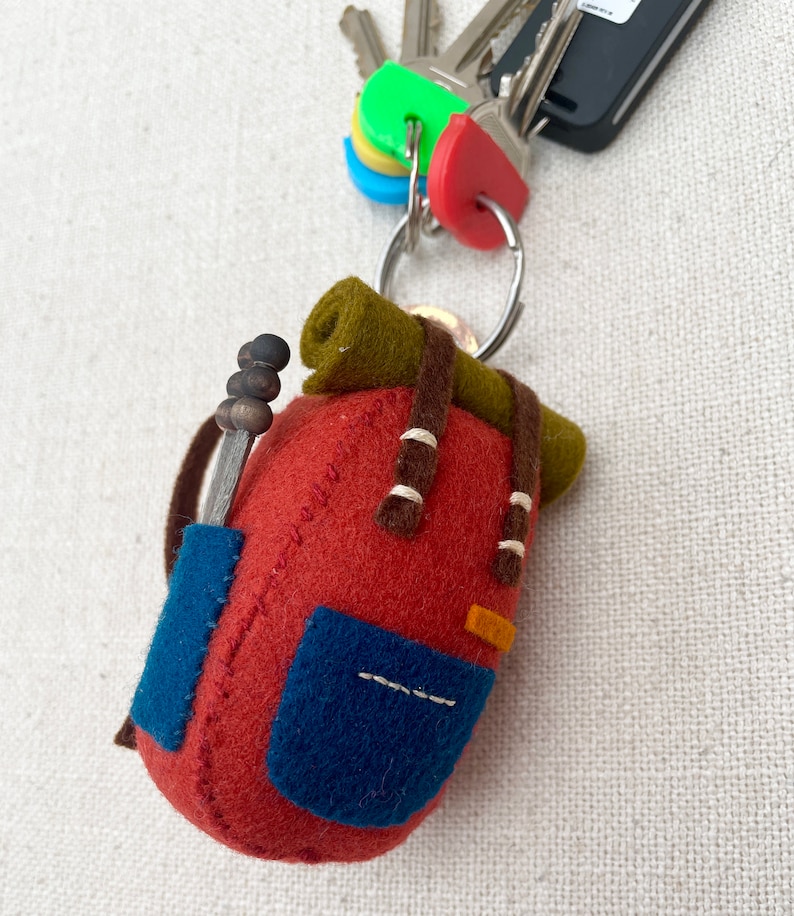 May include: A felt keyring shaped like a miniature backpack in rust red with a dark olive green bedroll on top, a blue front pocket, brown straps, and a small grey tool with three dark beads attached to the side, hanging from a metal ring with keys.