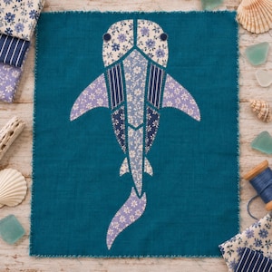 May include: A rectangular teal fabric panel featuring a central shark appliqué constructed from various blue and white floral and striped patterned fabric pieces, with visible stitching along the edges of the shark shape and the fabric panel.