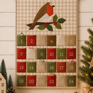 Pode incluir: Um calend&aacute;rio do advento em tecido com fundo de xadrez vichy bege e uma aplica&ccedil;&atilde;o de pisco-de-peito-ruivo num ramo de azevinho. O calend&aacute;rio tem vinte e quatro bolsos numerados em vermelho, verde e bege, suspensos por uma vara de madeira.
