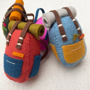 May include: A collection of four small, handcrafted felt backpacks in various colours including red, blue, green, and pink. Each miniature backpack features a rolled sleeping bag attached to the top, brown felt straps, and a small front pocket.