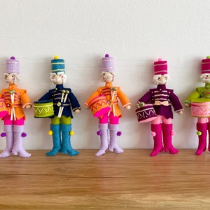 May include: Five felt toy drummers dressed in colourful uniforms with hats and holding drums. The drummers are standing in a row on a wooden surface.