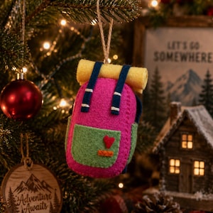 May include: A felt backpack ornament in bright pink with a light green front pocket, dark blue straps, and a yellow rolled sleeping bag on top. A small red heart and an orange rectangle are stitched onto the front pocket. It hangs by a twine loop.