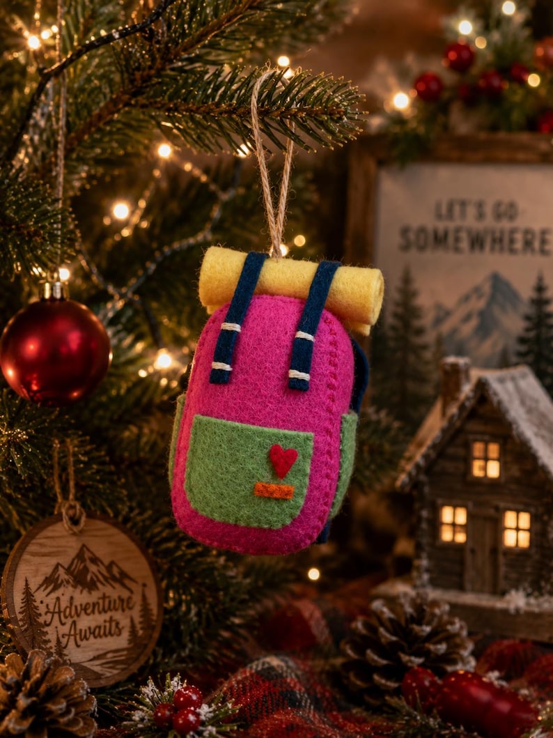 May include: A felt backpack ornament in bright pink with a light green front pocket, dark blue straps, and a yellow rolled sleeping bag on top. A small red heart and an orange rectangle are stitched onto the front pocket. It hangs by a twine loop.