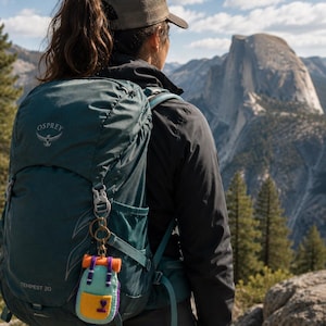 May include: A dark teal Osprey Tempest 20 backpack with a small, felted miniature backpack keychain attached to the strap. The miniature keychain features a light blue body, a yellow front pocket with a purple heart, and orange top straps.