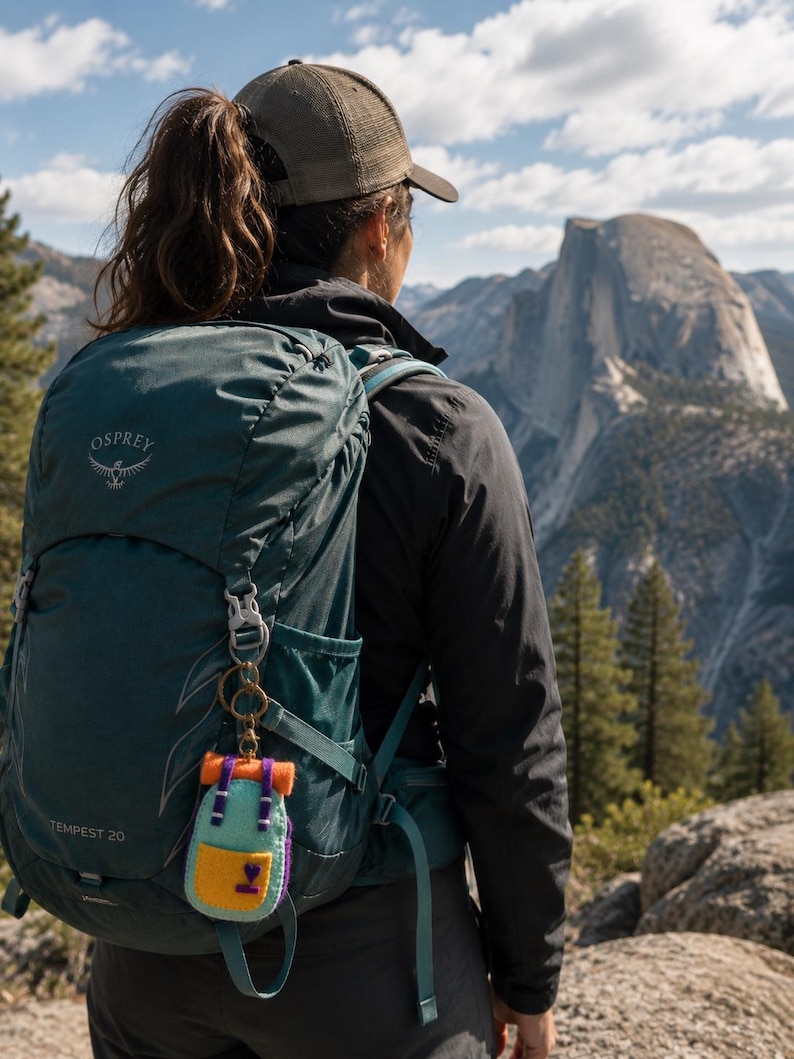May include: A dark teal Osprey Tempest 20 backpack with a small, felted miniature backpack keychain attached to the strap. The miniature keychain features a light blue body, a yellow front pocket with a purple heart, and orange top straps.