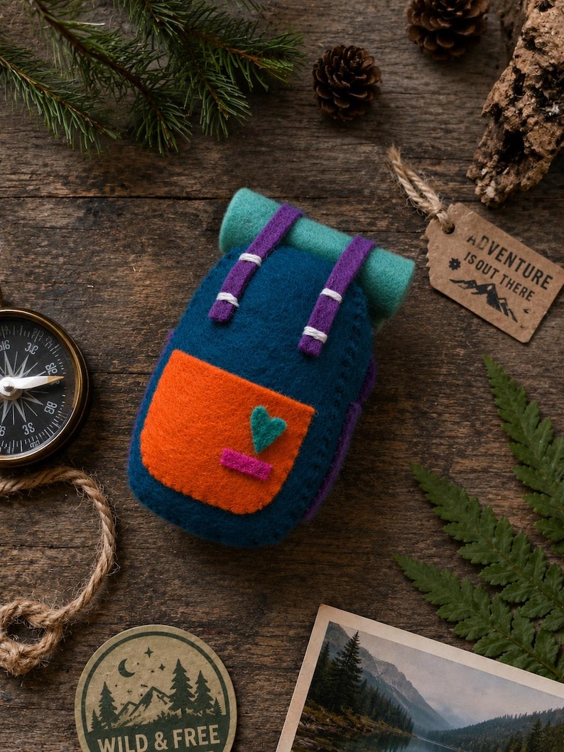 May include: A felt backpack ornament in deep blue with a bright orange front pocket, a teal top roll, and purple straps. The pocket features a small teal heart and a pink rectangle. The backpack is decorated with white stitching details.