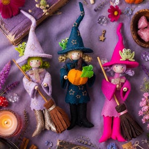May include: Three felt witch dolls in lavender, dark blue, and bright pink, each wearing a pointed hat with star embroidery and a small green frog. The dolls hold wooden broomsticks and feature painted faces, curly hair, and felt clothing.