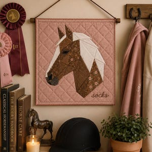 May include: A square quilted wall hanging featuring a geometric horse head applique in shades of brown and white with floral patterns on a dusty rose background. The word socks is embroidered in the bottom right corner in a cursive script font.