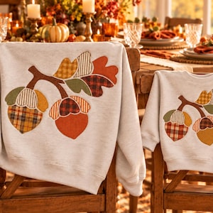 May include: Two light gray sweatshirts featuring a large applique of a brown branch with three acorns and leaves. The acorn caps and leaves are decorated with various plaid and solid fabric patterns in shades of orange, green, and brown.