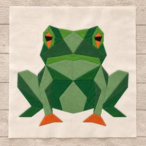 May include: A square fabric quilt block featuring a geometric frog design. The frog is constructed from various shades of green fabric pieces, with bright orange accents on the eyes and feet, all set against a solid off-white background.