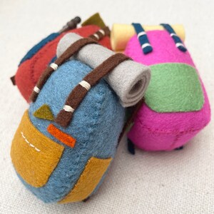 May include: Three miniature felt backpacks in blue, red, and pink. The blue backpack features a grey rolled sleeping bag, brown straps, and yellow pockets. The pink backpack has a green front pocket and blue straps. The red backpack is partially visible.