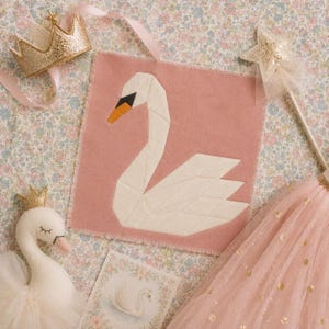 May include: A square dusty pink fabric wall hanging featuring a geometric white swan appliqué with a black and orange beak. The scene includes a gold glitter crown, a star-topped wand, a plush swan toy, and a pink tulle skirt with gold dots.