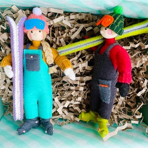 Puede incluir: Dos adornos de esquiadores de fieltro en una caja de regalo. Uno lleva un mono turquesa, un gorro rosa y gafas azules, sujetando esquís morados. El otro lleva una camisa roja, un peto negro y un gorro verde, llevando esquís amarillos.