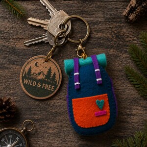 May include: A felt backpack keyring with a dark blue body, orange front pocket, teal top roll, and purple straps, attached to a round wooden charm engraved with a mountain scene and the text WILD & FREE, connected to a metal ring with keys.