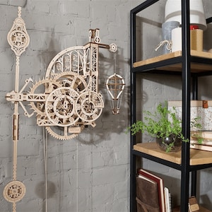 Könnte beinhalten: Ein Holz-Uhrenbausatz mit komplizierten Zahnrädern und einem Heißluftballon-Design. Die Uhr ist an einer grauen Backsteinwand neben einem schwarzen Metallregal montiert.