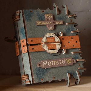 Può includere: Un libro blu-grigio con superficie testurizzata, dotato di una cinghia marrone al centro con una fibbia circolare con le scritte START e STOP. La copertina presenta targhe in legno con il testo The monster Book of MONSTERS e linguette laterali.