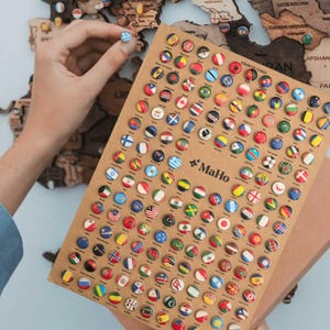 Può includere: Un foglio rettangolare di cartone color marrone chiaro con una griglia di numerose piccole spille circolari, ognuna delle quali mostra una diversa bandiera nazionale dai colori vivaci. Il testo MaHo è stampato in nero al centro del foglio.