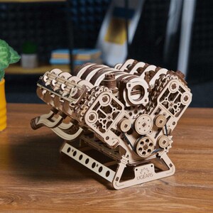 Könnte beinhalten: Ein mechanisches V8-Motormodell aus hell- und dunkelbraunem, lasergeschnittenem Sperrholz. Die komplexe Konstruktion zeigt sichtbare Zahnräder, Riemenscheiben, Gummibänder und eine Bodenplatte mit dem eingravierten Markennamen UGEARS.
