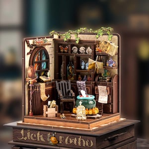 Può includere: Un diorama in miniatura in legno con una libreria marrone scuro, una piccola sedia nera e un calderone verde. La scena include una vetrata colorata, decorazioni a farfalla, una scopa, pergamene e una gabbia dorata. La base reca la scritta Lick to Potho.