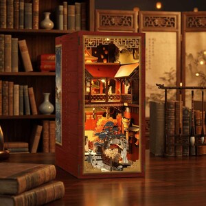 Könnte beinhalten: Ein rechteckiger Buchnischen-Einsatz aus Holz mit einer beleuchteten Miniaturszene eines traditionellen ostasiatischen Innenhofs mit roten Gebäuden, einer Steinbrücke, einem Baum und kleinen Figuren in einem dunkelroten Rahmen.