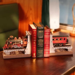 Könnte beinhalten: Ein dekoratives Buchstützen-Set mit einer schwarz-roten Dampflokomotive auf der einen und einem roten Personenwagen auf der anderen Seite, beide auf Schienen auf braunen Sockeln montiert, die drei Bücher mit rot-schwarzen Einbänden einrahmen.