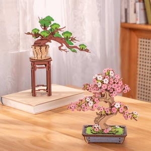 Könnte beinhalten: Zwei dekorative Bonsai-Baummodelle aus Holz. Einer zeigt grünes Laub auf einem hohen braunen Ständer, während der andere rosa und weiße Kirschblüten in einem rechteckigen blauen Topf präsentiert. Beide bestehen aus geschichtetem Lasercut-Holz.