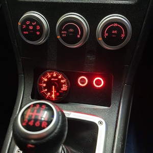 May include: A black car interior with a red and silver gear shifter, a red gauge with a black face, and three silver control knobs with red lights. The knobs are labelled "AUTO", "AUTO OFF", and "A/C".